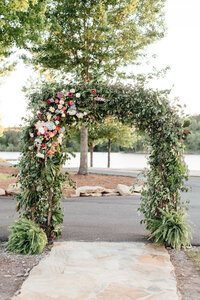 An archway covered in vines and greenery with pink and white flowers in the corner of the arch way