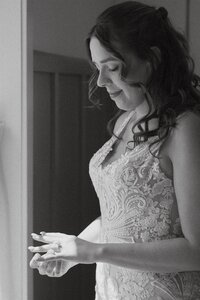 Bride standing near window in elegant lace wedding dress, gently admiring her ring with a soft smile — captured in black and white captured by a Destination Elopement Photographer at Stone Spring Farms, Pennsylvania.