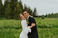 Bridals shoot in Rexburg, Idaho. A bride and groom hold each other, their cheeks touching. Both are smiling at the camera. They are in a grassy field with pine trees behind them.
