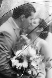 black and white image of bride and groom kissing underneath a clear umbrella in the rain