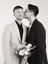 Black and white film portrait of two grooms in a studio setting, one holding a bouquet and smiling as his partner kisses him on the cheek — captured by a film wedding photographer.