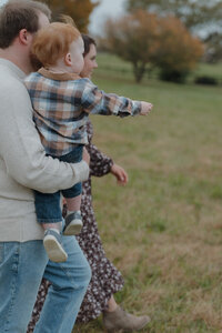 A family walks in a field in Limestone, TN. The father is carrying their young son while the son points at something. There are trees in the background.