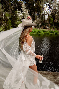 Boho bride wearing a wide-brim hat and flowing veil, walking near water in Florida during a playful, western-inspired bridal session.