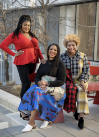 Jae Merchant in a red top and black pants standing near Jasmine Hudson who is wearing a black top with blue and gold skirt. Sitting near Jasmine is  Madia Willis who is wearing a plaid patchwork dress thats black, grey, gold and red.