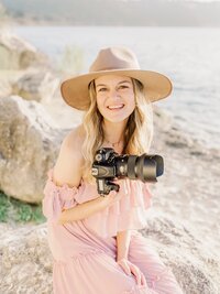 Karina Danielle Photography is smiling, holding wedding camera sitting on a rock on Lake Dallas.