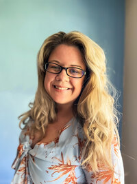 Smiling blonde woman wearing glasses and blue patterned dress smiles at camera