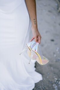 A bride in a white dress holding her wedding shoe while walking on sand.