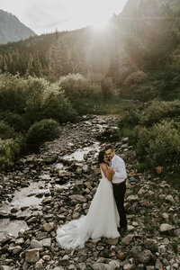 Bride and groom share a romantic moment by a rocky creek with sun rays peeking through pines in Breckenridge, Colorado.