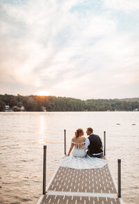 A bride and groom sitting on the edge of a dock over looking the sunset over the lake with their backs to the camera.