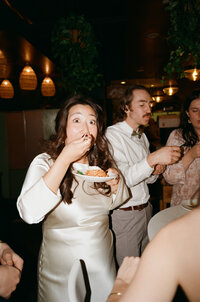 Candid film photo of a bride in a satin gown taking a playful bite of food during her wedding reception — captured by a film wedding photographer at Castillo Holliday Photo + Film.