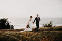 A bride and groom walking down a trail on the edge of the beach
