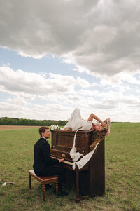 Groom plays a piano in a field while bride lays atop\