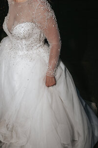 Close-up view of an elegant white beaded wedding gown featuring sheer long sleeves and a voluminous tulle ball gown skirt, showcased against a dark background for a dramatic and luxurious bridal fashion portrait.