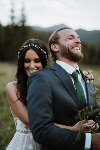 Bride hugs groom from behind in a mountain meadow, both laughing during wedding photos in Breckenridge, Colorado.