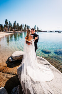 Bride and groom embracing on the rocky shoreline of Lake Tahoe, surrounded by crystal-clear blue water and stunning mountain views.