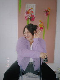 A person with dark purple hair styled in a loose updo sits on a clear acrylic chair, laughing with eyes closed while flashing a peace sign. They wear a lilac cardigan over a white top, black pants, and gold jewelry, holding a small vintage film camera. The image has a retro, direct-flash aesthetic, with a pink floral-decorated wall in the background. A timestamp in the corner adds to the nostalgic feel.
