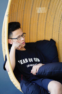 A young, masculine presenting teen sits in a wicker chair, leaning into the back, which extends up above their head. They are framed by one pillow behind them and the other in their lap, with one arm wrapped around it. They rest their head on their free, right arm, looking past the camera.