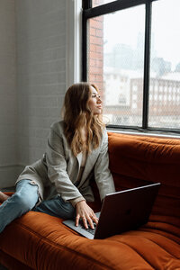 female entrepreneur sitting on an orange couch with her laptop out stares out the window