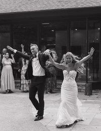 A joyful couple in wedding attire enters a venue, holding hands and raising arms in celebration. Guests cheer in the background. Black and white photo.