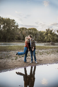 A couple kissing by a small body of water with a reflection visible, surrounded by trees and a cloudy sky.