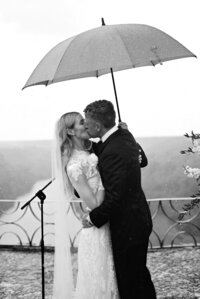 Bride and groom sharing a kiss under an umbrella during a rainy wedding ceremony — timeless black and white moment captured by Asia Pimentel Photography.