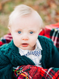 Baby in green sweater sits in a silver metal tub with red plaid blanket, looking up.
