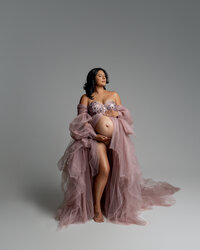 Pregnant woman wearing a flowing mauve tulle gown stands barefoot in a studio, cradling her baby bump with both hands and looking upward with a serene expression.
Photograph by Yucaipa maternity photographer Kaitlyn Dawn Photography.