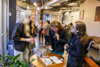 A woman with long hair and a cream-colored shirt is pouring a drink from a carton at a counter filled with various pastries and baked goods. Another woman stands nearby in the background, holding a cup. The scene, reminiscent of a relaxed networking event, takes place in a well-lit room in Seattle.