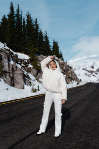 Artist in an all-white outfit standing confidently on a scenic snowy road – bold winter brand session by Mewphotovideo