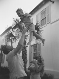 Black and white image of dad tossing son in the air while mom holding baby smiles at them