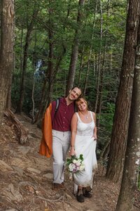 A bride and groom stand on a hiking trail surrounded by lush green pine trees with a waterfall off in the distant background. The bride is in a flowy wedding dress with a long veil and the groom is wearing tan pants with an orange leather jacket. 
