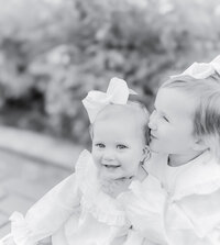 Black and white portrait of two young sisters dressed in ruffled outfits with matching bows, as the older sister leans in to give a sweet kiss. Their joyful expressions and close bond reflect the warmth and timeless beauty of childhood, captured by a Birmingham family photographer.