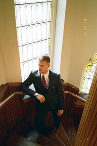 Groom leaning on a curved wooden staircase railing in a suit.