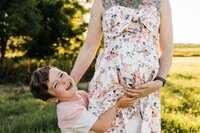 Young boy hugging a pregnant woman's belly, both smiling, in a sunny park setting suitable for family photography.