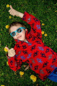 Boy wearing sunglasses lying on the grass surrounded by dandelions giving the thumbs up sign