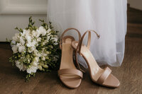 The brids shoes stand besides the buquet on the floor.