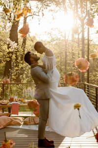 A groom is picking up his bride. The photo is backlit by the sun, and their are colorful flowers in the foreground