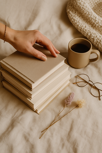 A flat lay image featuring a woman’s hand reaching for a layered stack of books on a cream-toned background, styled with soft textiles and cozy neutral tones — perfect for book lovers, self-development, and personal growth inspiration.