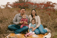 Two parents sitting on a blanket in the grass each holding a small child 