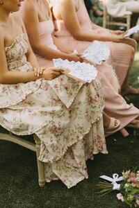Details of women in pink and floral dresses sitting in wooden chairs holding white fans in a lawn