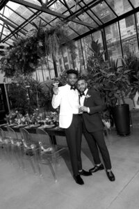Queer grooms in front of a modern wedding tablescape in a greenhouse venue in Milwaukee