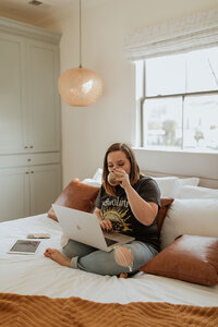 A woman sits cross-legged on a bed with rust-colored pillows, using a laptop and sipping from a mug. An iPad is beside her as she explores a white label web design course, with daylight streaming in through the window behind her.