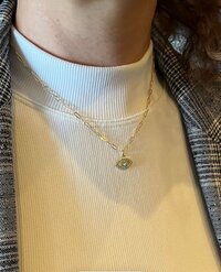 Gold charm necklace with an evil eye pendant worn over a white mock neck top.