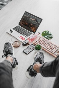 A top down view of a woman's legs in dark jeans and patterned black shoes. On the floor is a laptop, stylized keyboard, phone, and other aesthetic items like floral, drink, keys.