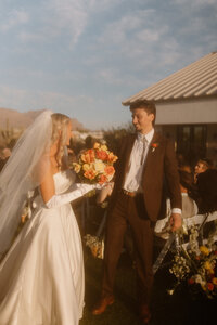 old money western wedding ceremony, the couple is leaving their cermony after having just gotten married. You see the back of the bride with her classic wedding gown, elbow length sleefves, and yellow and orange wedding bouquet. The groom looks excitedly at his bride. He's wearing a dark brown suit with a bolo tie.