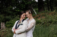Two brides embrace on their wedding day. Their foreheads are close to one another, and their eyes are closed with emotion.