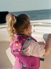 A young child in a vibrant, star-patterned life jacket gazes out over a boat's edge towards the water. The scene is serene and playful.