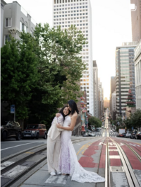 Queer brides at sunrise in San Francisco holding each other, one in a custom purple floral suit and the other in Quill gown by Claire Pettibone