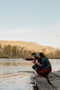 A husband and wife and standing on the edge of a lake. They are holding their son and pointing into the distance.