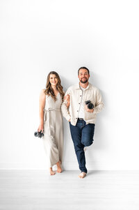 Clients of Bostron Design Studio, Kala holds Reg's arm as they both smile with their cameras. They are in a white studio.